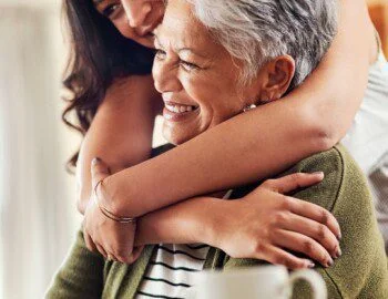Woman hugs older woman from behind, both smiling warmly.