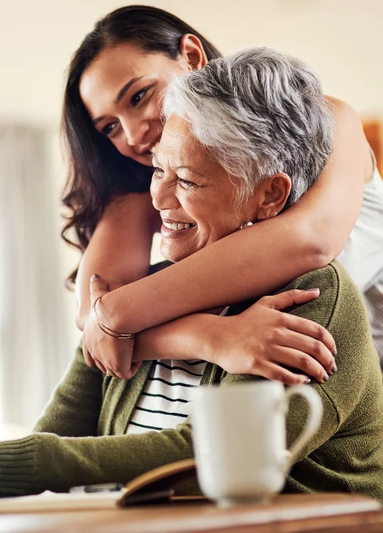 Woman hugs older woman from behind, both smiling warmly.