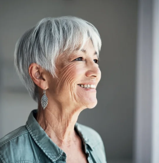 Smiling elderly person with short gray hair and leaf-shaped earring in profile view.