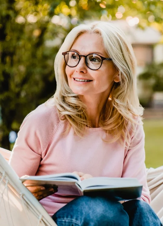 Woman in glasses sitting outdoors, smiling, holding a book.
