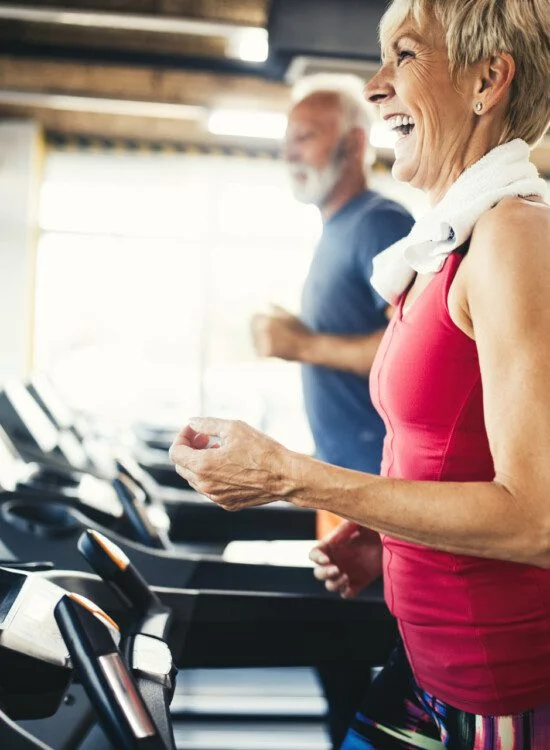 Older adults running on treadmills at the gym, smiling and exercising.