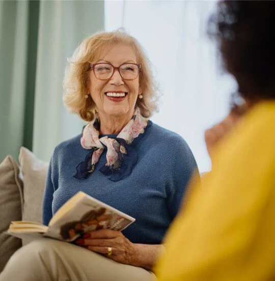 Smiling woman holding book, talking with someone in yellow shirt.