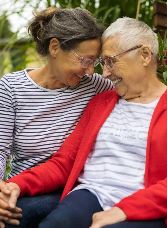Two women smiling and embracing, surrounded by plants outdoors.