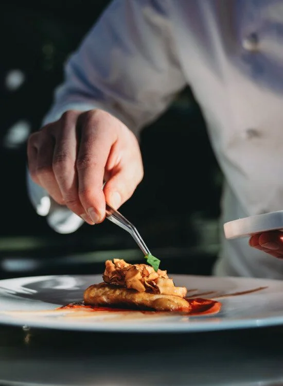 Chef garnishing a plated dish with precision using tweezers in a kitchen setting.