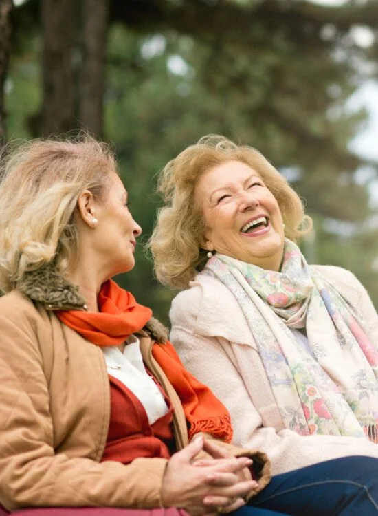 Two elderly women laughing together on a park bench outdoors.