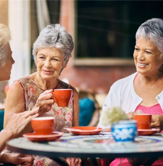 Three elderly women enjoy coffee and conversation at a patio table.