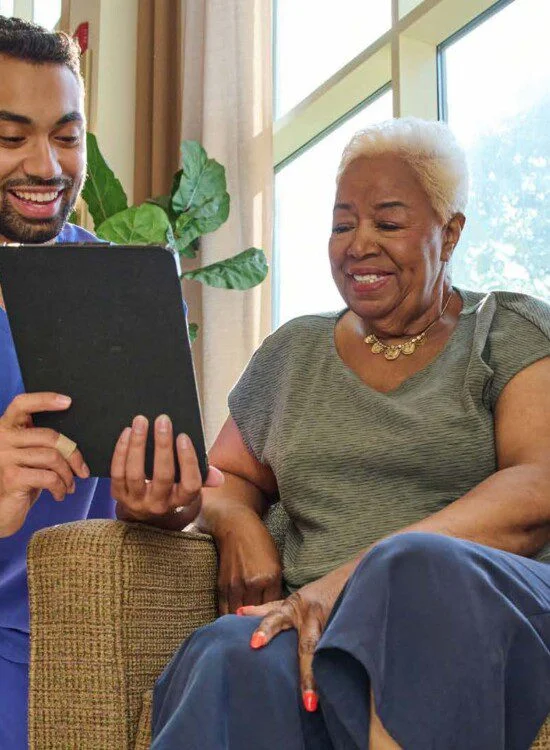 Caretaker shows tablet to smiling elderly woman in a bright room.