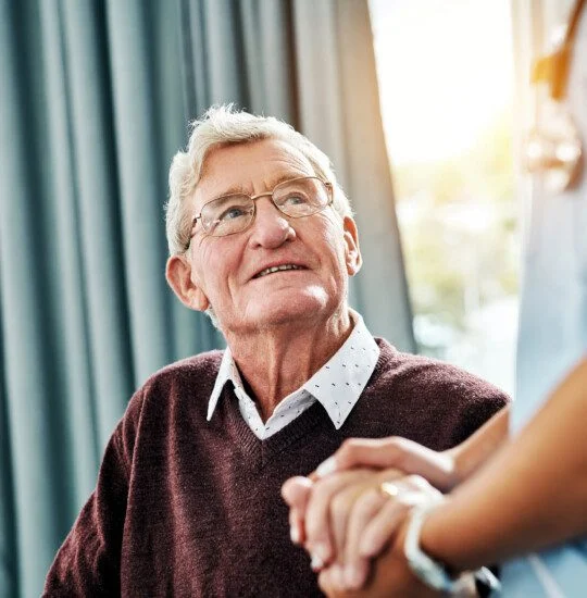 Elderly man smiling at a caregiver holding his hand indoors.
