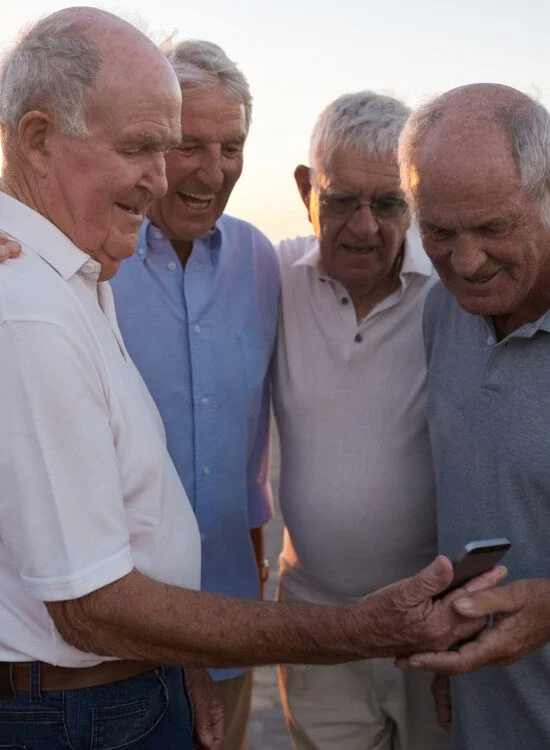 Four elderly men smiling and looking at a smartphone outdoors.