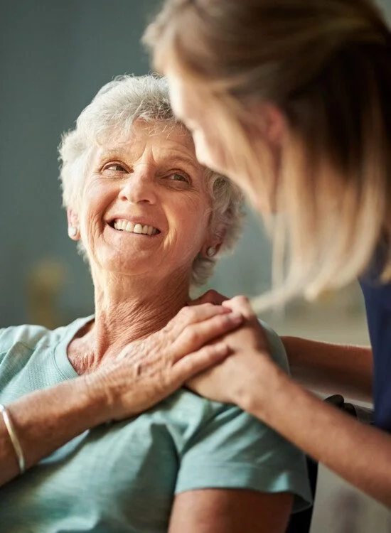 Elderly woman smiling as caregiver holds her shoulder, showing warmth and care.