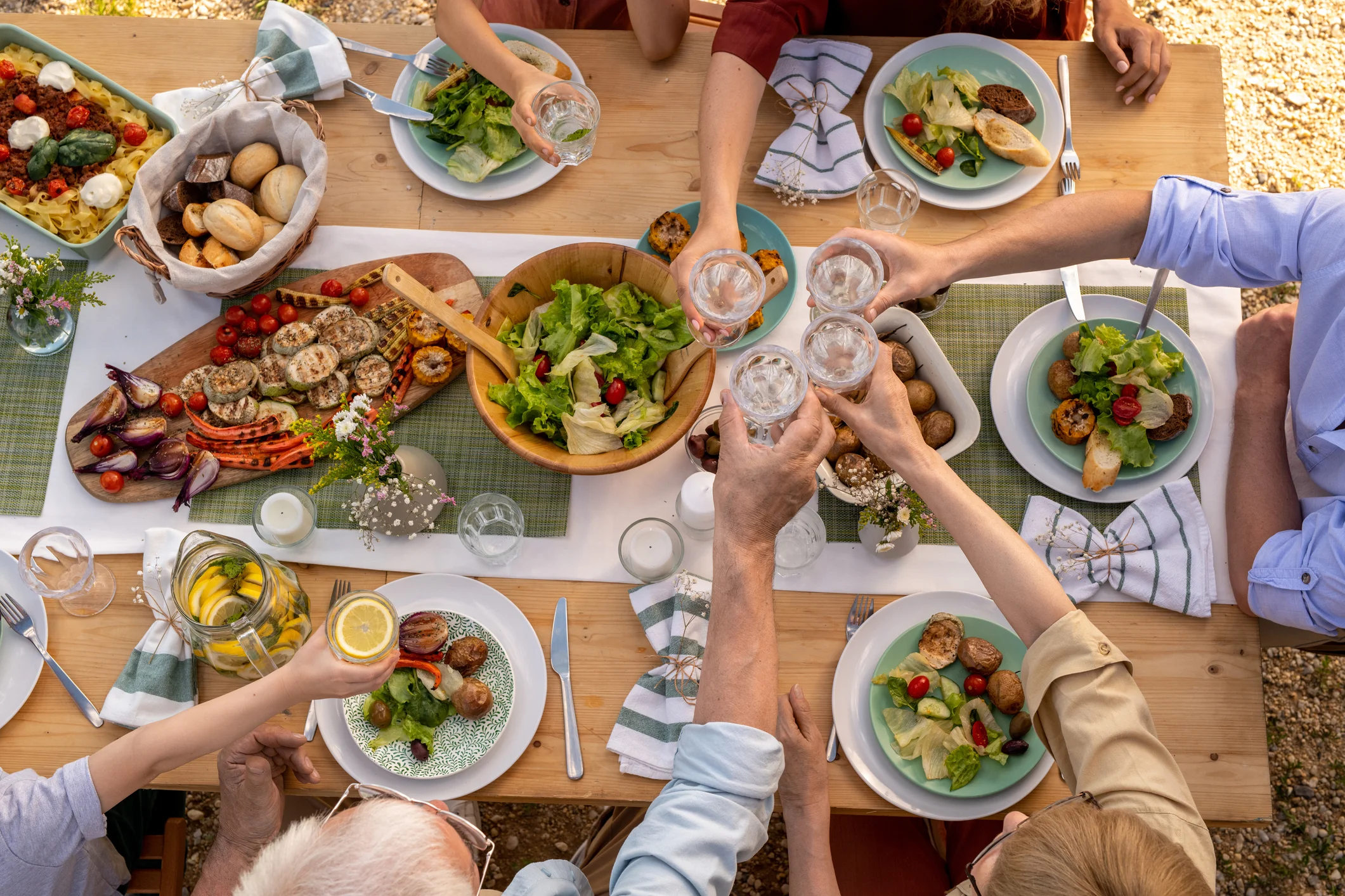 People toasting over an outdoor meal with salads, bread, and roasted vegetables.