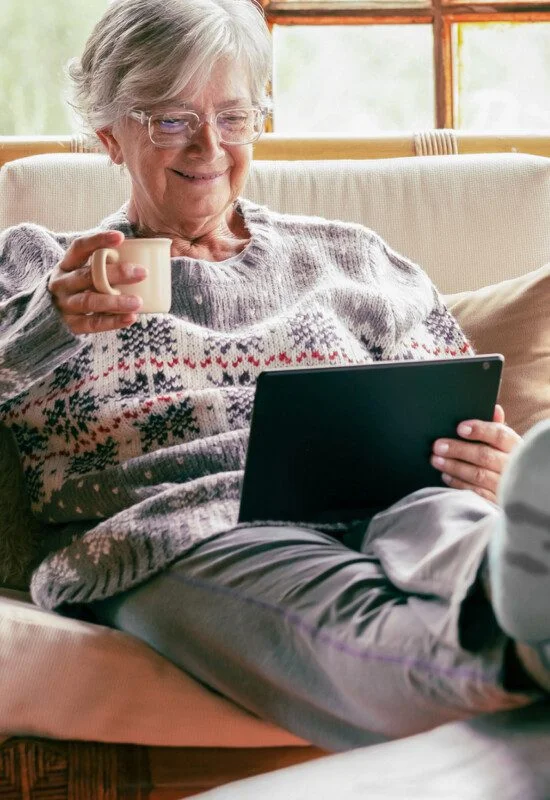 Elderly person relaxing with tablet and coffee mug on a sofa, wearing a cozy sweater.