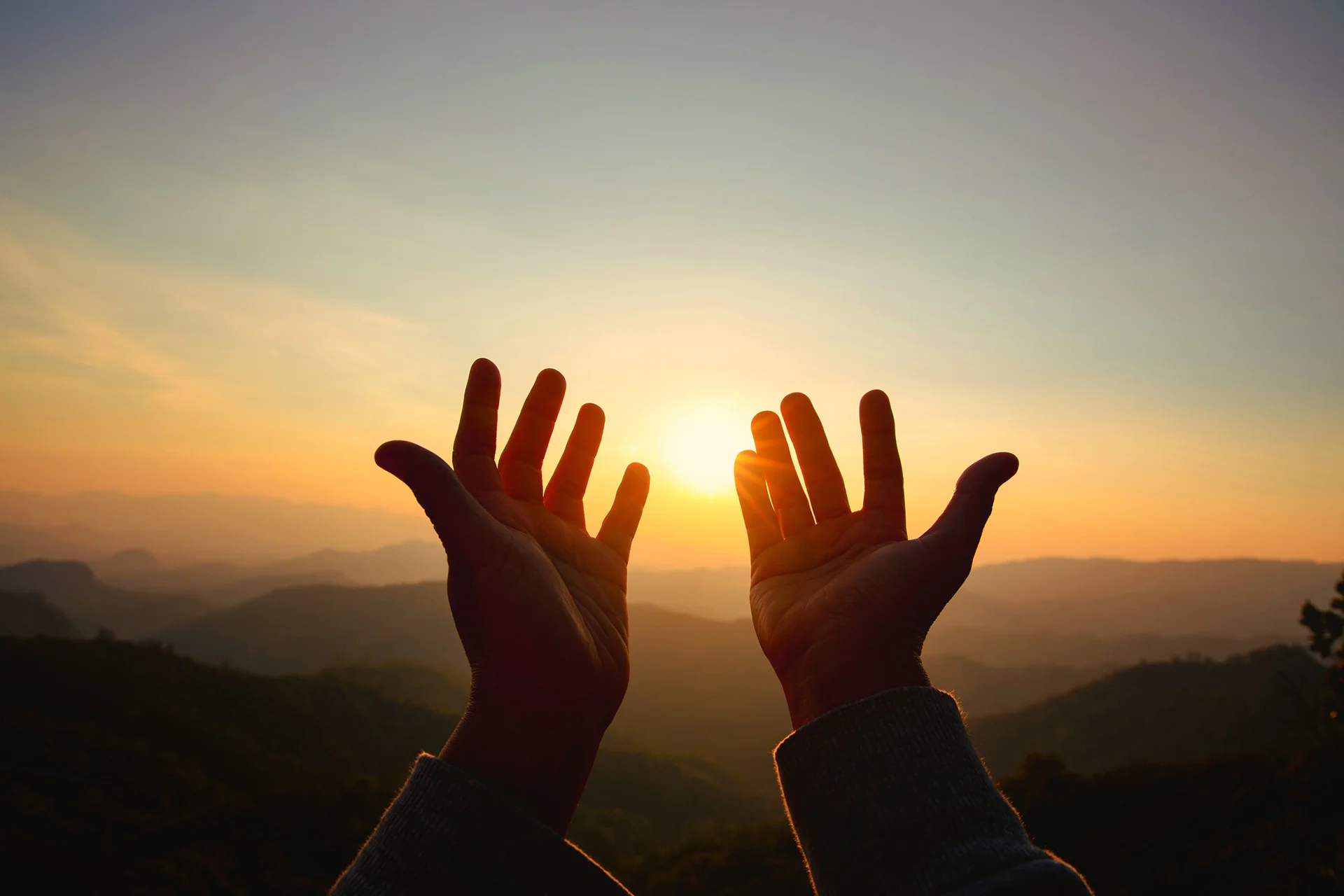 Hands raised towards a sunset over distant mountains.