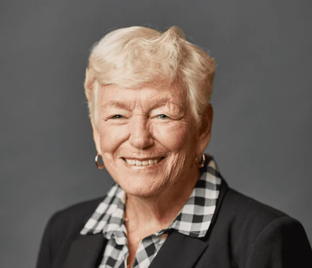 Smiling person with short hair in checkered shirt and blazer against gray background.
