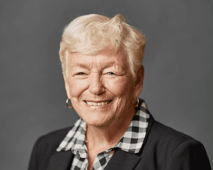 Smiling person with short hair in checkered shirt and blazer against gray background.