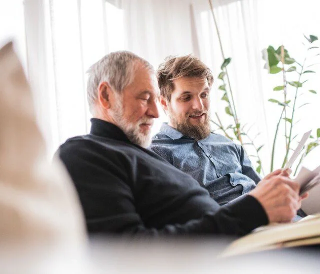 Two men sitting on a couch, reading and smiling together, with plants in the background.