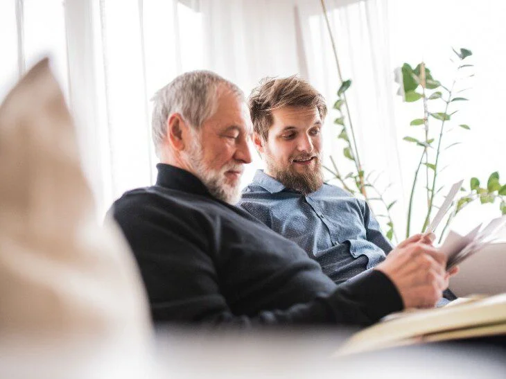 Two men sitting on a couch, reading and smiling together, with plants in the background.