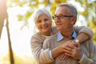 Older couple smiling in a sunny park, woman embracing the man from behind.
