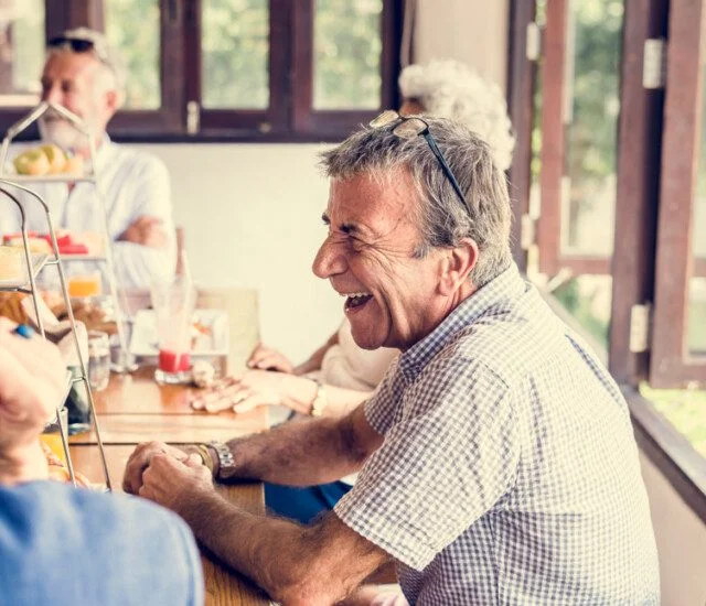 Older adults laughing at a table with food and drinks in a bright room.