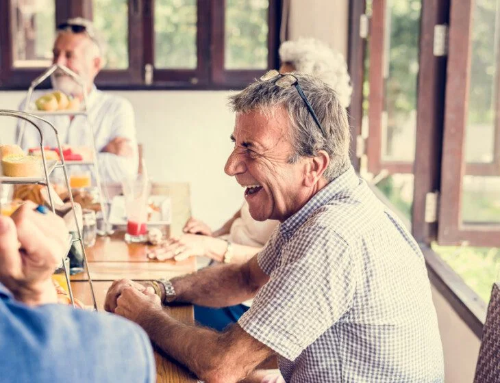 Older adults laughing at a table with food and drinks in a bright room.