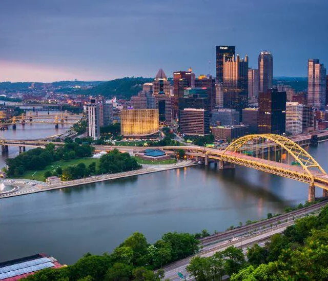 City skyline with yellow bridges over rivers and a riverside park at dusk.