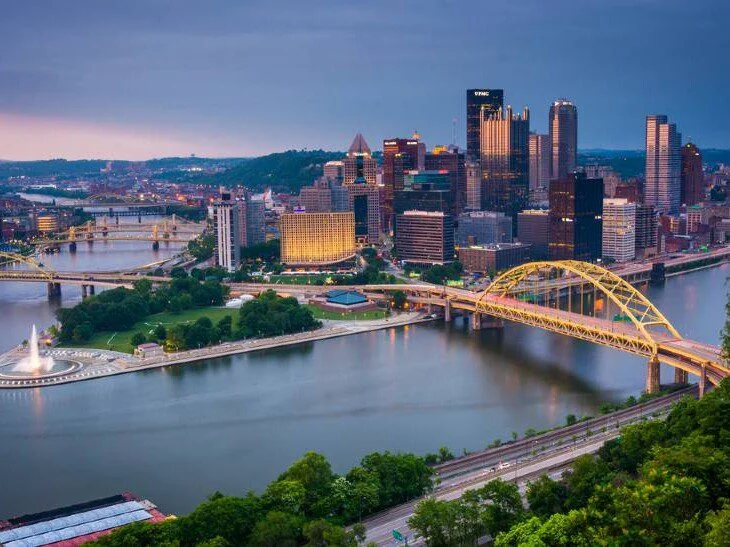 City skyline with yellow bridges over rivers and a riverside park at dusk.