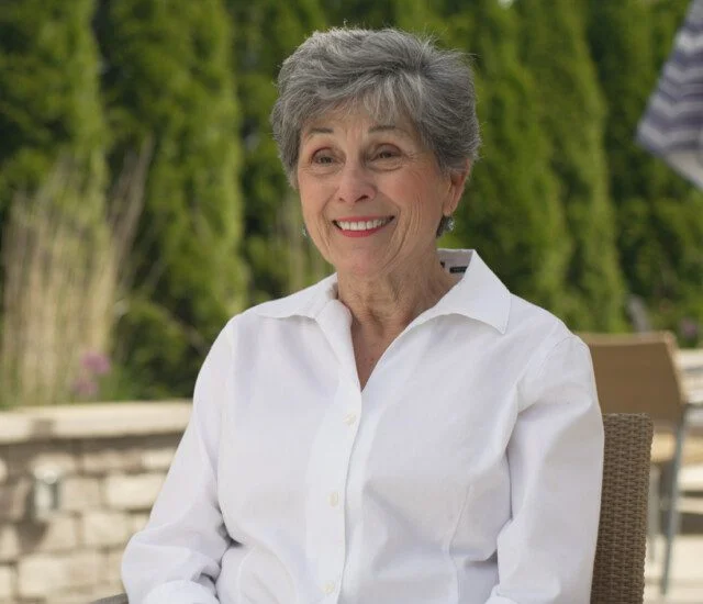 Smiling woman in a white shirt sitting outdoors with greenery in the background.