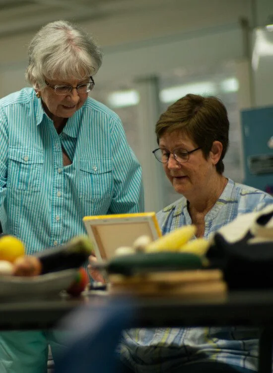Two women discussing a document in a kitchen setting with fruits on the table.