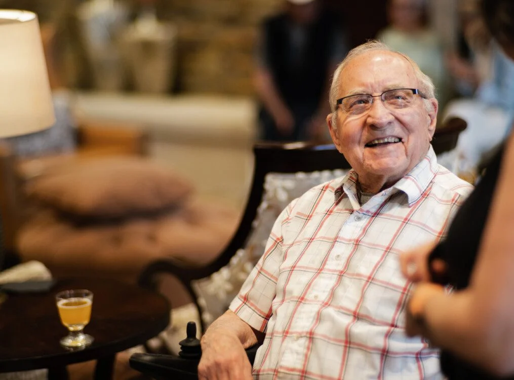 Elderly man smiling while speaking with someone in a cozy room setting.