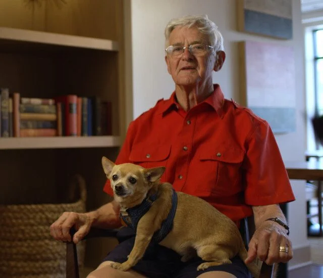Elderly person in red shirt sitting, holding a small dog indoors.