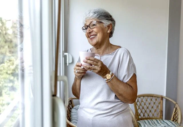 Woman with glasses smiling and holding a mug while looking out the window on a sunny day.