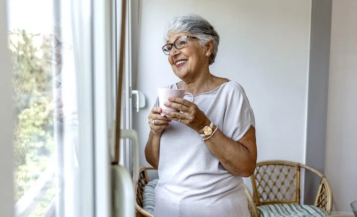 Woman with glasses smiling and holding a mug while looking out the window on a sunny day.