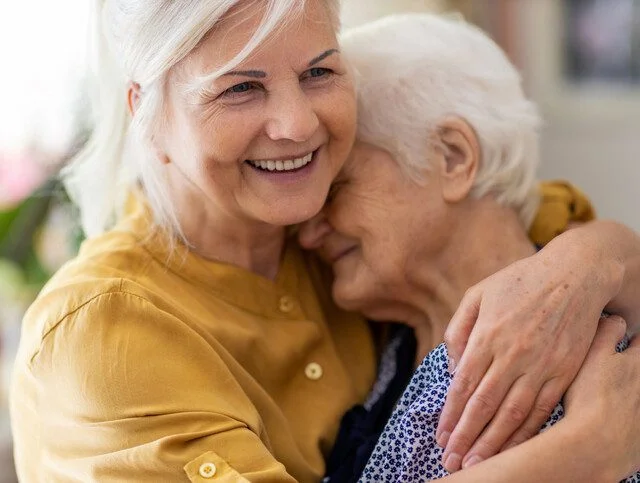Two elderly women hugging and smiling warmly indoors.