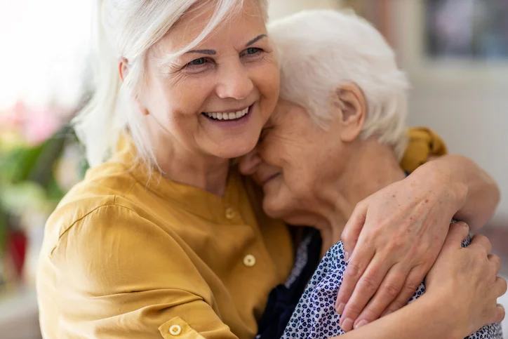 Two elderly women hugging and smiling warmly indoors.