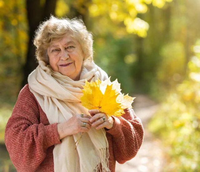 Elderly woman in scarf holding yellow leaves in sunny park.