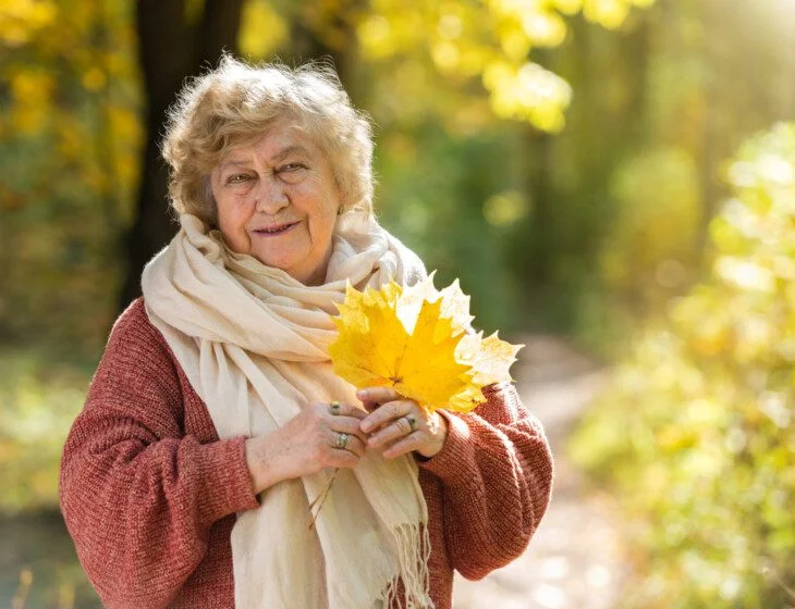 Elderly woman in scarf holding yellow leaves in sunny park.