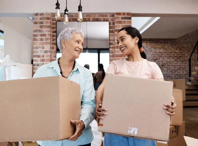 Two women smiling and holding boxes in a modern room with brick walls.