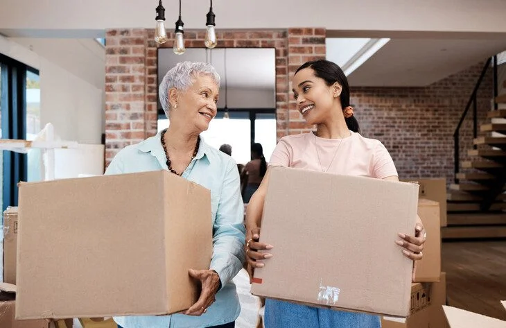 Two women smiling and holding boxes in a modern room with brick walls.