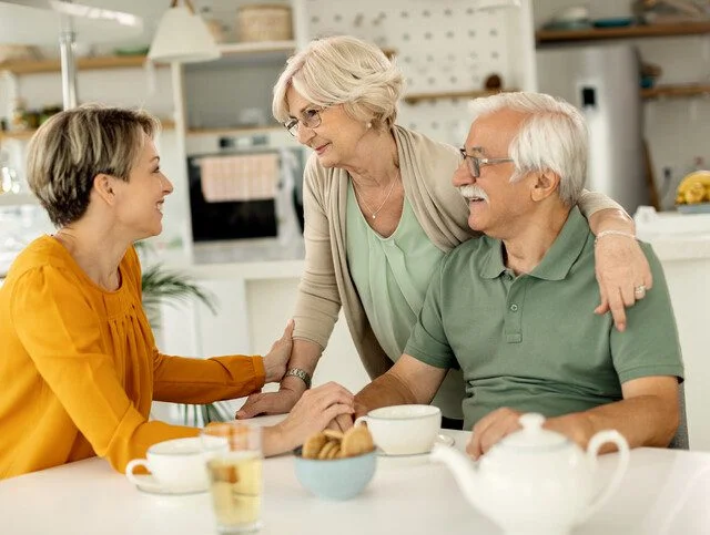 Older couple and woman smiling, sitting at kitchen table with tea and snacks.