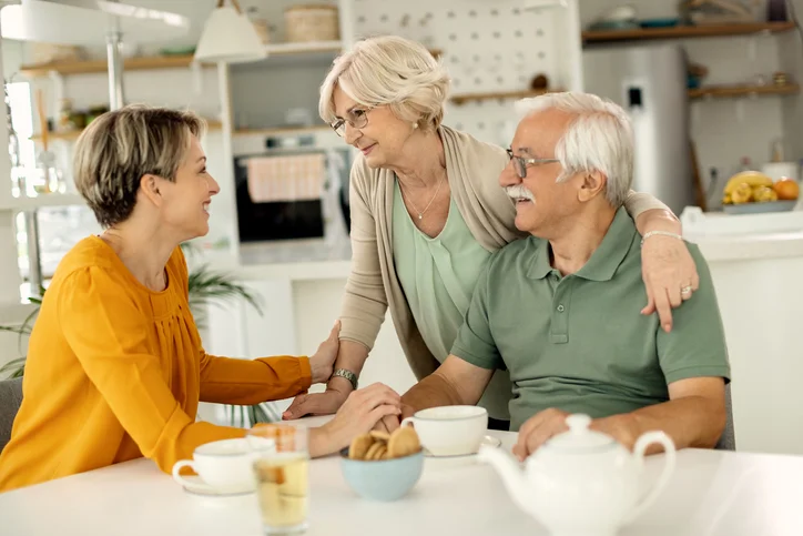 Older couple and woman smiling, sitting at kitchen table with tea and snacks.