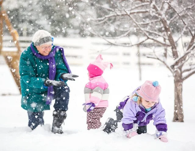 Grandparent and two children play in the snow, surrounded by trees and snowfall.