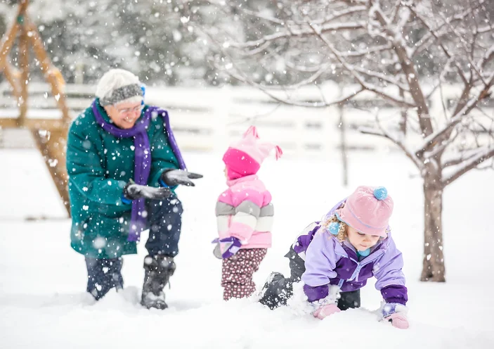 Grandparent and two children play in the snow, surrounded by trees and snowfall.