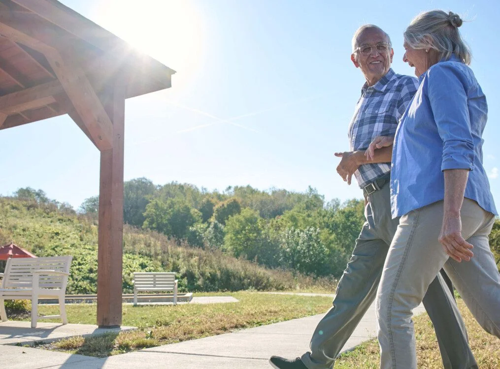 Elderly couple walking outside near a gazebo on a sunny day.