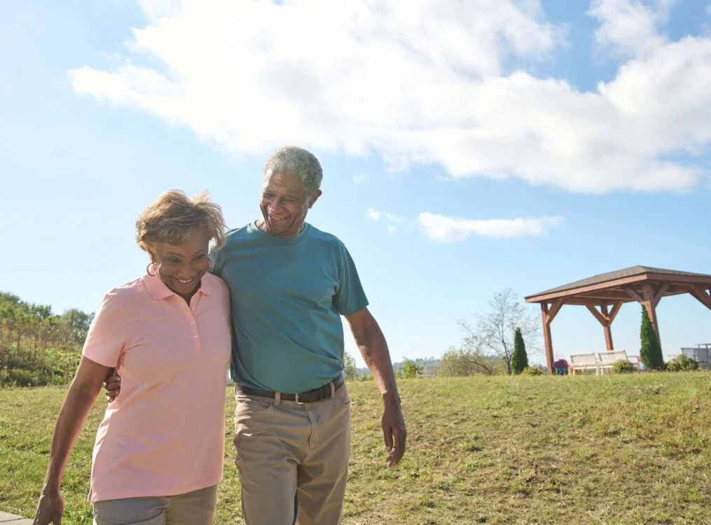 Elderly couple walking together on a sunny day near a wooden gazebo.