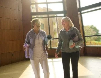 FVSH Selects R2 LR (1) (1) Page 13 Image 0001 Two women laughing, holding yoga mats and water bottles in a sunlit room.