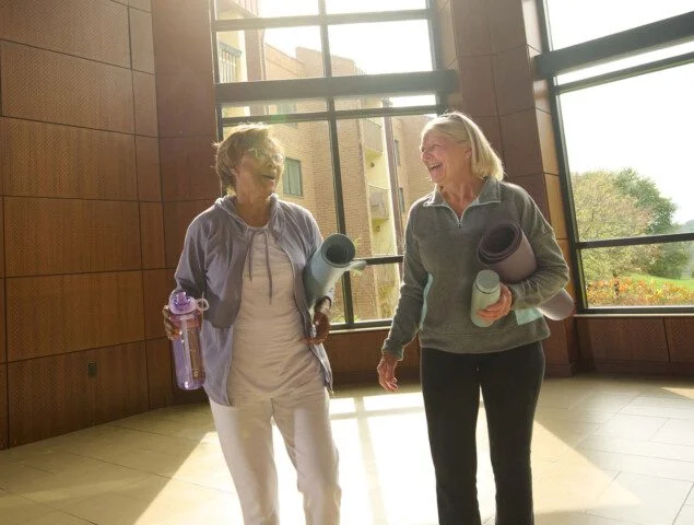 Two women laughing, holding yoga mats and water bottles in a sunlit room.