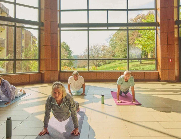 Older adults doing yoga indoors with large windows and garden view.