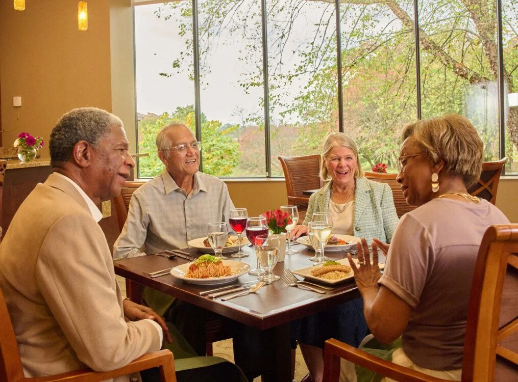 Four people dining together, smiling in a well-lit restaurant with large windows.