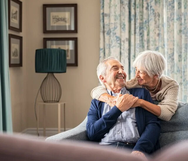 Elderly couple smiling, sitting on a sofa in a cozy, well-decorated living room.
