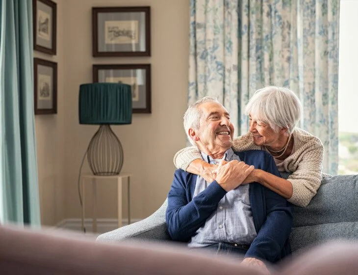 Elderly couple smiling, sitting on a sofa in a cozy, well-decorated living room.